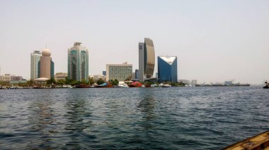 Limana yanaşmış çeşitli gökdelenler ve teknelerin yer aldığı bir liman boyunca uzanan modern bir şehrin manzarası. Gökyüzü açık, güneşli bir günü yansıtıyor..