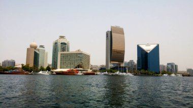 Rıhtım boyunca uzanan modern bir şehrin panoramik görüntüsü, ön planda teknelerin kenetlendiği çeşitli gökdelenleri ve binaları gösteriyor..