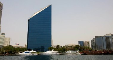 Benzersiz üçgen şekilli modern bir gökdelen, rıhtıma yakın lüks yatları ve tekneleri olan mavi camları yansıtıyor. Gökyüzünde çeşitli binalar ve palmiye ağaçları yer alıyor..