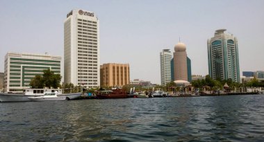 Nehir boyunca modern gökdelenleri ve geleneksel ahşap tekneleri gösteren bir rıhtım manzarası. Ufuk çizgisi, ön planda yeşillik bulunan mimari tarzların bir karışımını içerir..