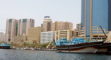 Arka planda modern gökdelenlerle kenetlenmiş geleneksel ahşap teknelerle dolu hareketli bir liman manzarası eski ve yeni mimarinin bir karışımını sergiliyor..