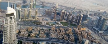 Uzun gökdelenlerin, mimari tarzların ve bir nehrin karışımının yer aldığı modern bir şehir manzarası. Ön planda alçak binaları olan yoğun bir alan gösterilirken, arka planda yüksek binalar ve çöl manzarası ön plana çıkıyor..