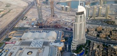 Gökdelenlerin, büyük bir alışveriş merkezinin ve bir otoyolun yer aldığı modern bir şehir manzarası. Sahne, kentsel mimari ve inşaat alanlarının bir karışımını yakalıyor ve arka planda su birimleri görülüyor..