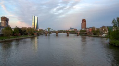 Her iki tarafında modern binalar olan bir nehrin manzarası. Bir köprü nehri geçer, akşam ışığını yansıtır. Gökyüzü parçalı bulutlu, sakin bir atmosfer yaratıyor..