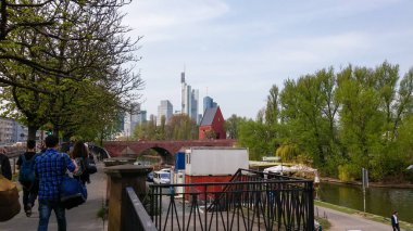 Nehir kenarında yürüyüş yapan insanların manzaralı bir manzarası. Arka planda, modern gökdelenler tarihi kırmızı bir binanın üzerine yükselir ve yeşil ağaçları yemyeşil ederek, doğa ve kentsel mimarinin bir karışımını yaratır..