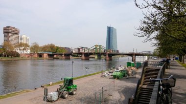 Arka planda modern bir bina olan nehir manzarası, nehri geçen bir köprü ve nehir kıyısında inşaat ekipmanları. Ağaçlar kaldırıma dizilmiş, ve insanlar manzaranın tadını çıkarırken görülüyor..