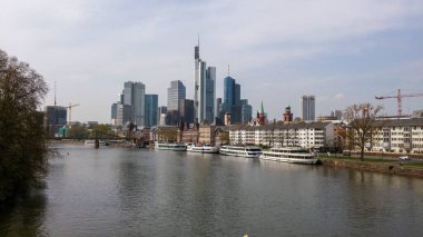 Nehir boyunca uzanan Frankfurt gökdelenleri ve tarihi binaları gösteren manzaralı bir manzara. Tekneler rıhtıma yanaştı, nehir kıyısında ağaçlar var..