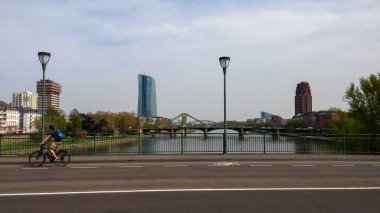 Bir bisikletçi nehir manzaralı ve arka planda modern binalar olan bir köprü boyunca seyahat eder. Sahnede açık bir gökyüzü altında kentsel mimari ve yeşillik karışımı yer alıyor..