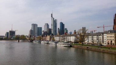 Frankfurt, Almanya 'nın panoramik manzarası nehir kenarındaki tarihi binaların yanında modern gökdelenleri gösteriyor. Ufuk çizgisi, kentsel gelişimi gösteren vinç ve inşaata sahip..
