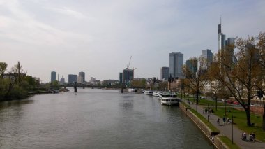 Arka planında modern gökdelenler olan bir nehrin manzarası, nehir kenarında sıralanan ağaçlar ve kıyı boyunca boş zamanların keyfini çıkaran insanlar..