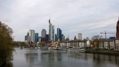 Nehir boyunca uzanan modern bir şehrin manzarası. Nehir kıyısında yüksek gökdelenler ve tarihi binalar var. Üzerinde bulutlu bir gökyüzü var..