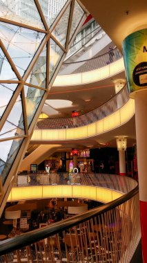 Modern bir alışveriş merkezinin içinde, yumuşak ışıklandırmayla aydınlatılmış birden fazla katlı spiral bir merdiven var. Dizayn cam ve metal elementleri içeriyor, çağdaş bir atmosfer yaratıyor..