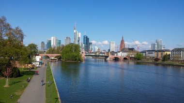 Nehir boyunca uzanan Frankfurt gökdelenlerinin ve tarihsel mimarinin manzarası. Gökyüzü açık ve mavi, nehir boyunca uzanan bir patika var..