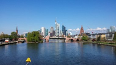 Modern gökdelenlerin, tarihi binaların ve bir nehrin yer aldığı Frankfurt 'un gökyüzünün manzarası. Gökyüzü birkaç bulutla berrak ve nehir kıyıları boyunca yeşillik görülebilir..
