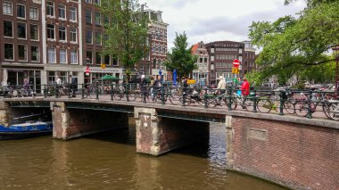 Amsterdam 'da bisikletçiler ve yayalarla dolu yoğun bir köprü. Bisikletler tırabzan boyunca park edilmiş ve kanal çevredeki binaları ve ağaçları yansıtıyor. Atmosfer canlı ve kentsel..