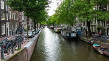 Amsterdam 'da ağaçlar ve geleneksel Hollanda binalarıyla dolu pitoresk bir kanal sahnesi. Tekneler su boyunca demirli ve bisikletler yakınlarda park halinde, sakin bir kentsel atmosfer yaratıyorlar..