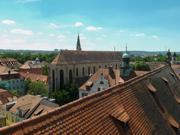 Çatıları olan panoramik bir manzara, uzun çan kulesi olan bir kilise ve yeşil ağaçlar. Gökyüzü parlak mavi tüylü beyaz bulutlar, dingin bir atmosfer yaratıyor..