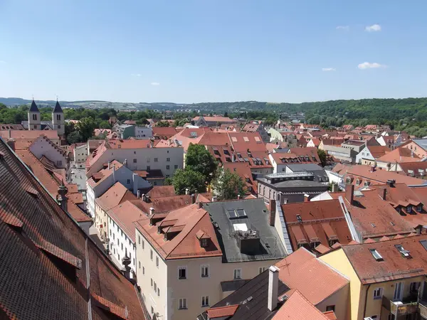 Kırmızı kiremitli çatıları, arka planda yeşil tepeleri ve açık mavi gökyüzü olan büyüleyici bir Avrupa kasabasının panoramik görüntüsü. Sahne, geleneksel mimarinin ve huzurlu manzaranın özünü yakalıyor..