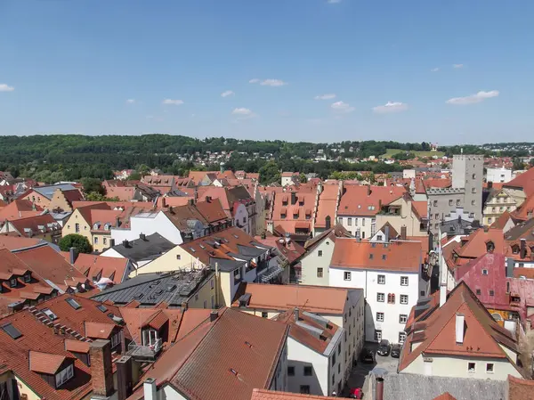 Açık mavi gökyüzünün altında kırmızı kiremitli çatıları olan antika bir kasabanın panoramik görüntüsü. Manzara, arka planda geleneksel binalar ve yeşillik karışımından oluşuyor..