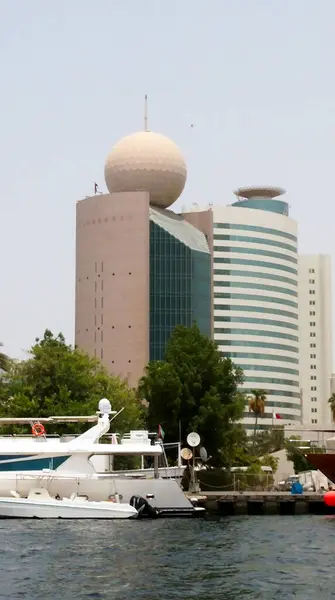 Tepesinde küre şeklinde bir kubbe olan modern bir bina, rıhtıma yanaşmış tekneleri olan bir liman. Mimaride yeşil alanlarla çevrili cam ve beton elementler yer alıyor..