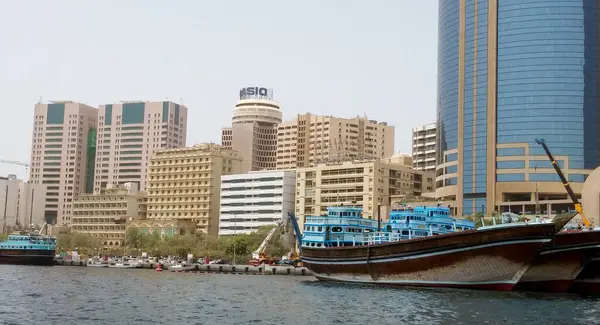 Arka planda modern gökdelenlerle kenetlenmiş geleneksel ahşap teknelerle dolu hareketli bir liman manzarası eski ve yeni mimarinin bir karışımını sergiliyor..