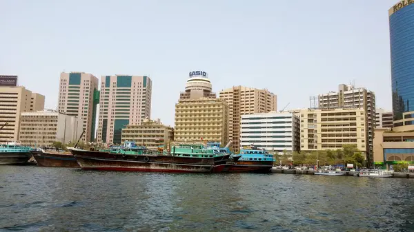 Arka planda modern gökdelenler olan bir rıhtım boyunca demirlenmiş geleneksel ahşap teknelerin manzarası. Sahne, hareketli bir kentsel alanda eski ve yeni mimari arasındaki zıtlığı yakalıyor..