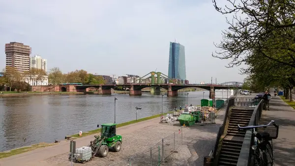 Arka planda modern bir bina olan nehir manzarası, nehri geçen bir köprü ve nehir kıyısında inşaat ekipmanları. Ağaçlar kaldırıma dizilmiş, ve insanlar manzaranın tadını çıkarırken görülüyor..