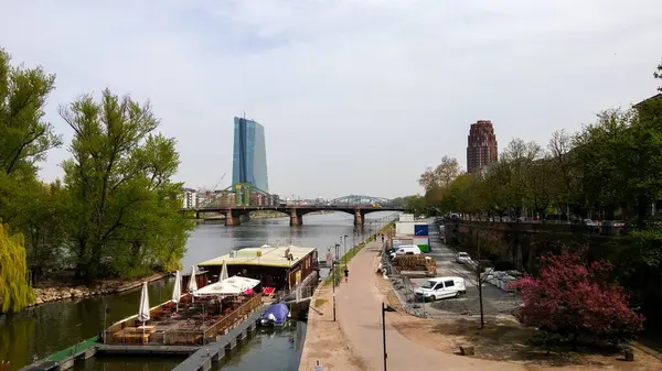 Yürüyüş yolu olan bir nehir manzarası, kenetlenmiş tekneler ve arka planda modern binalar. Ağaçlar yolu çiziyor, ve bir köprü nehri geçiyor, sakin bir şehir manzarası yaratıyor..