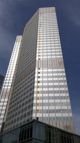 Gökyüzünü yansıtan cam bir cephesi olan uzun, modern bir gökdelen. Bina yatay ve dikey çizgileri olan eşsiz bir tasarıma sahiptir ve bazı pencereler kısmen açıktır. Ön planda cam panelleri olan daha alçak bir yapı var..