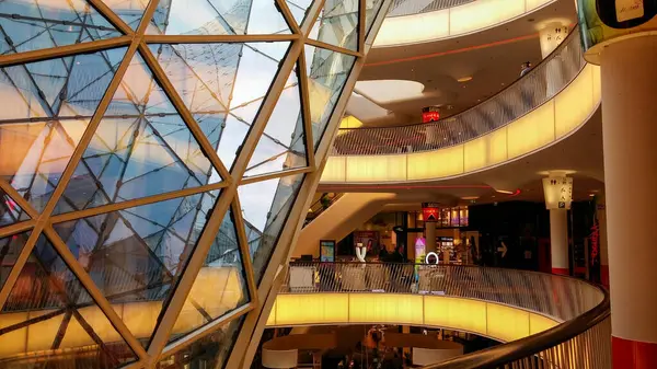 Geometrik cam yapısı olan modern bir alışveriş merkezi. Aydınlatılmış korkulukları ve arka planda müşterileri olan çeşitli katlar..