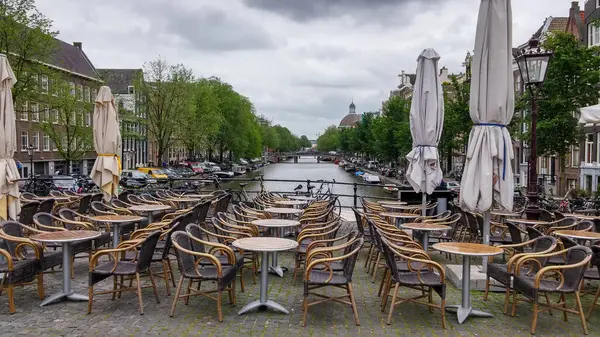 Amsterdam 'da boş kafeterya masaları ve sandalyeleri olan bir kanal manzarası. Bulutlu gökyüzü karamsar bir atmosfer eklerken yemyeşil ağaçlar su yoluna diziliyor.