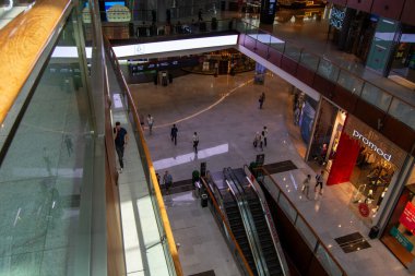 Yürüyen merdivenleri, cam korkulukları ve etrafta dolaşan müşterileri olan geniş bir alışveriş merkezi. Arka planda çeşitli depolarla aydınlık.