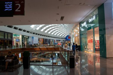 Modern mimarisi, büyük cam pencereleri ve parlak bir atmosferi olan geniş bir alışveriş merkezi. Müşteriler, arka planda çeşitli mağazalar bulunan cilalı zemin boyunca yürürler..