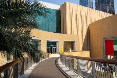 Büyük cam pencereleri ve dokulu altın bir cephesi olan bir binaya uzanan eğri ahşap bir geçidi olan modern bir mimari yapı. Ön planda bir palmiye ağacı görülebiliyor ve sahne yüksek katlı bir şehir ortamında geçiyor.