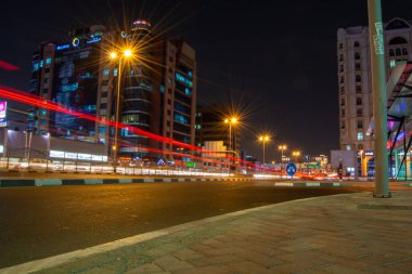 Yoldan geçen araçların ışık yollarıyla dolu yoğun bir yola sahip bir gece şehri. Sokak lambaları ve neon ışıklarıyla aydınlatılmış uzun binalar. Sahne, karanlık çöktükten sonra kentsel yaşamın canlı enerjisini yakalıyor..