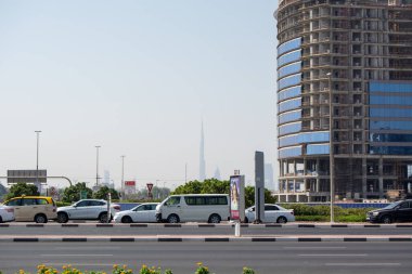 Yol boyunca park etmiş birkaç arabanın bulunduğu işlek bir şehir caddesi. Arka planda, modern bir şehir manzarasına işaret eden kısmen görünen uzun bir gökdelen var. Ön planda yeşillik ve inşaat binaları yer alıyor, devam eden gelişmeler sergileniyor