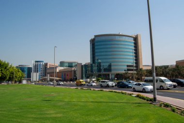 Yüksek binaların, açık mavi gökyüzünün ve ön planda iyi korunmuş yeşil çimlerin bulunduğu modern bir şehir manzarası. Araçlar yol boyunca park halinde, kalabalık bir şehir ortamı sergiliyorlar..