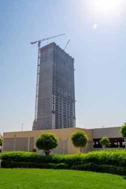 Yapım aşamasında olan uzun bir inşaat kulesi, iskelesi ve vinci var. Bina, arka planda açık mavi gökyüzü ve ön planda yeşil çimlerle kısmen tamamlanmıştır..