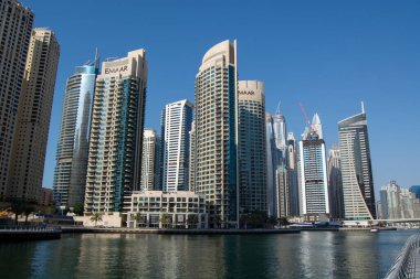 Dubai 'deki modern gökdelenlerin gökyüzü manzarası mimari stil ve tasarımların bir karışımını sergiliyor. Binalar, ön planda su bulunan açık mavi gökyüzüne karşı güneş ışığını yansıtıyor..