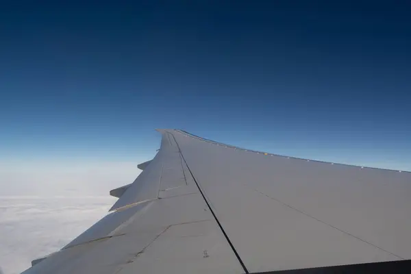 Bir uçak penceresinden hava görüntüsü, kanadı açık mavi gökyüzüne ve aşağıdaki tüylü beyaz bulutlara karşı gösteriyor..