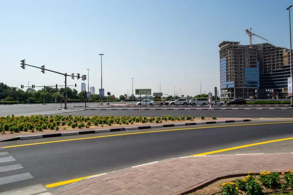 Trafik ışıkları, bakımlı bir yol ve çiçek tarhı olan işlek bir şehir kavşağı. Arka planda, bir inşaat alanı ve modern binalar açık mavi gökyüzünün altında görülebilir..