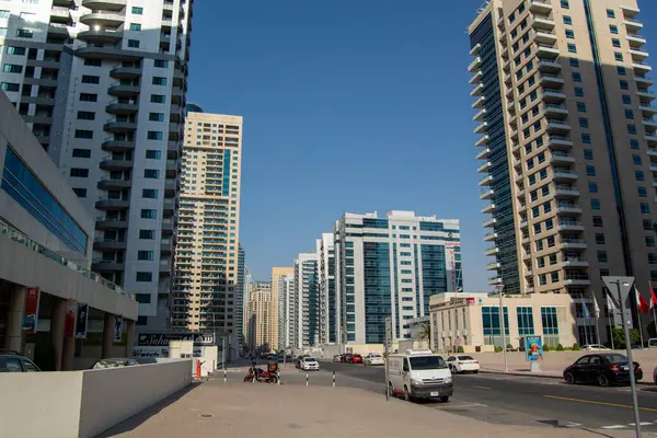 Açık mavi gökyüzüne sahip, yüksek konut ve ticari binaların yer aldığı modern bir şehir manzarası. Sokaklar taşıt ve yayalarla dolu, hareketli bir şehir atmosferi sergiliyorlar..