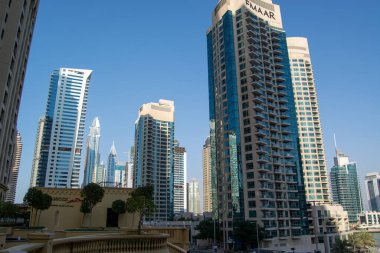 Bir şehirdeki modern gökdelenlerin görüntüsü, açık mavi gökyüzü altında mimari stiller ve cam cephelerin karışımını sergiliyor..