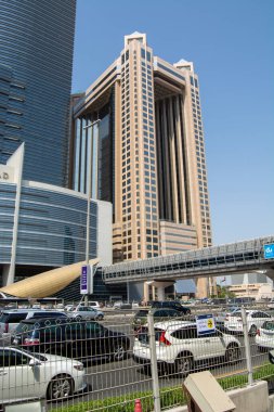 Uzun gökdelenler ve yaya köprüsünün yer aldığı modern bir şehir manzarası. Ön planda çeşitli arabalarla dolu yoğun bir park alanı gösterilirken, arka planda eşsiz mimari tasarıma sahip önemli bir bina ön plana çıkıyor..