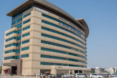 'AL MIDEA MARKET PLACE' yazan ve büyük cam pencereleri olan modern çok katlı bir bina. Yapı, önünde araçlar park edilmiş, açık mavi bir gökyüzüne karşı kurulmuştur..