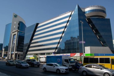 Gökyüzünü yansıtan büyük cam pencereli, benzersiz açısal tasarımı olan modern bir ticari bina. Bina, önünde çeşitli araçlar bulunan bir kentsel alanda yer almaktadır..