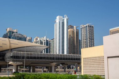 Dubai 'de açık mavi gökyüzü olan yüksek binaların karışımından oluşan modern bir şehir silueti. Ön planda çağdaş bir mimari yapı ve yeşillik yer alıyor..