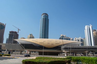 Pürüzsüz, kıvrımlı bir çatısı ve büyük cam pencereleri olan, gelecekçi bir tren istasyonuna benzeyen modern bir mimari yapı. Gökdelenleri çevreleyen açık mavi gökyüzü şehir manzarasını güçlendirir.