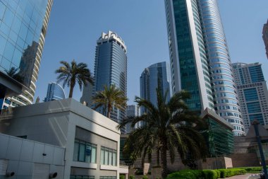 Açık mavi gökyüzünün altında yüksek gökdelenler ve palmiye ağaçları bulunan modern bir şehir manzarası. Binaların yansıtıcı cam cepheleri var, mimari tarzların bir karışımını sergiliyorlar..