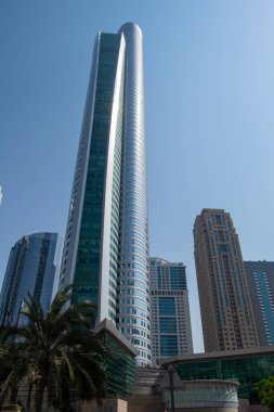 Açık mavi gökyüzünün altında, diğer binalar ve palmiye ağaçları tarafından çevrelenmiş, parlak cam cepheli, uzun, modern bir gökdelen..
